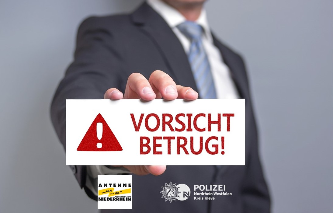 das Bild zeigt einen Mann im Anzug, der ein weißes Schild mit dem Text "Vorsicht Betrug" in die Kamera hält. Außerdem ist auch ein rot-weiß "Achtung" Zeichen auf dem Schild. Unter dem Schild sind die Logos von Antenne Niederrhein und der KPB Kleve abgebildet. 