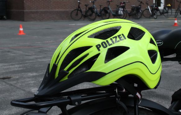 ein reflektierender Helm, mit der Aufschrift Polizei, liegt auf dem Gepäckträger eines Pedeles. Im Hintergrund stehen mehrere Pylonen sowie weitere Pedelec an einer Wand. 