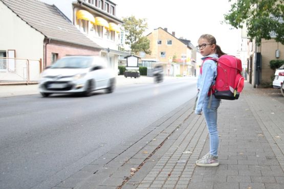 während ein junges Mädchen am Straßenrand steht und dabei einen roten Rucksack trägt, fährt auf der Straße ein Pkw und dahinter ein Motorrad. Die beiden Verkehrsteilnehmenden sind verschwommen dargestellt. Die Häuserfronten erinnern an eine belebte Kleinstadt.